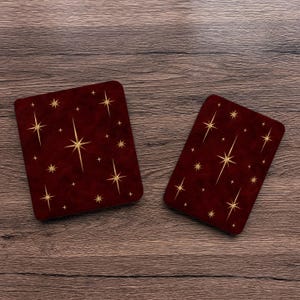 Pode incluir: Dois porta-copos retangulares com um fundo vermelho bordô profundo, apresentando um padrão repetitivo de estrelas douradas de quatro pontas e pequenos brilhos cintilantes espalhados pela superfície de cada peça.