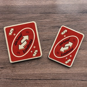 May include: Two red coasters with the text "spread kindness" in a white oval. The coasters have a white border and feature white arrows and starburst designs. The coasters are on a wooden surface.