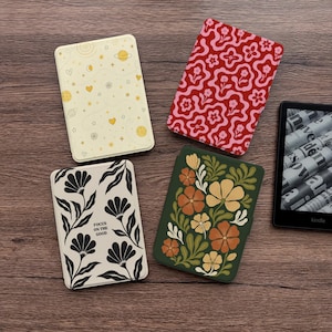 Puede incluir: Cuatro fundas decorativas para Kindle con varios diseños. Una funda es de color crema con un patrón celestial de planetas, estrellas y corazones. Otra es roja con un patrón floral rosa. Una tercera es crema con ilustraciones florales negras y el texto "FOCUS ON THE GOOD". La última es verde con flores naranjas y amarillas.