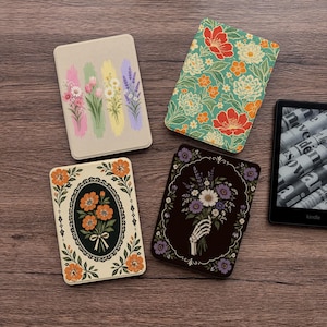 May include: Four rectangular e-reader cases with distinct designs: one with vertical floral brushstrokes, one with a teal floral pattern, one with an orange flower bouquet in an oval frame, and one with a skeletal hand holding a bouquet.