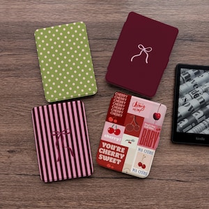May include: Four rectangular e-reader cases and one black e-reader. One case is green with white polka dots, one is solid maroon with a white bow, one is pink and maroon striped with a bow, and one features a cherry-themed collage with text.
