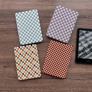 May include: Four rectangular cases with checkered patterns in blue, purple, red, and multicolored squares. A Kindle e-reader is visible on the right side of the image, displaying a newspaper. The cases are on a wooden surface.
