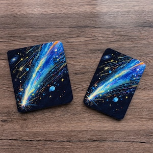 May include: Two rectangular coasters featuring a vibrant space scene with a bright yellow and blue comet streaking across a dark navy background filled with scattered stars, distant galaxies, and small blue planets in a cosmic art style.
