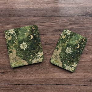 May include: Two rectangular coasters featuring a dark green botanical print with white flowers, golden suns, crescent moons, and scattered stars. The design includes intricate foliage and small butterflies, creating a detailed celestial and nature-inspired pattern on both items.