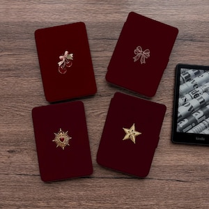 May include: Four rectangular dark red e-reader cases with rounded corners. One features a cherry charm with a bow, one a gold bow outline, one a gold sunburst heart emblem, and one a gold star. A black Kindle e-reader is visible on the right.