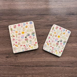 May include: Two off-white rectangular cases with a colourful floral and whimsical design. The cases are covered in illustrations of flowers, rainbows, teddy bears, and other decorative elements. The cases are on a brown wooden surface.