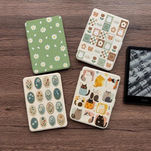 Può includere: Quattro custodie per Kindle con vari design sono esposte su una superficie di legno. Le custodie presentano motivi come margherite su verde, ovali floreali, una griglia con orsi e conigli e gatti dei cartoni animati. È visibile anche un dispositivo Kindle.