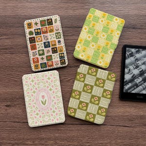 May include: Three rectangular e-reader cases with floral patterns and a black Kindle e-reader. One case features a grid of colorful flower squares, another has a yellow and green checkered daisy print, and the third has a pink tulip oval.
