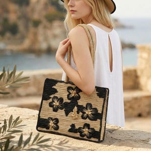 Puede incluir: Un bolso tote rectangular con un diseño tejido. El bolso presenta un fondo beige natural con detalles florales negros. Las asas y el ribete son de color beige a juego. El bolso lo lleva una persona que lleva un vestido blanco.