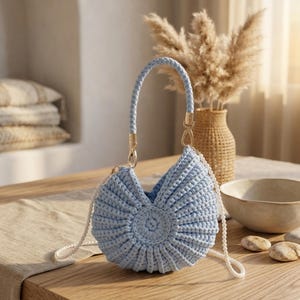 May include: A light blue, handmade crochet bag with a unique shell-like design. The bag features a braided handle and gold-colored hardware. A delicate white beaded strap is also attached. The bag is displayed on a wooden table, showcasing its craftsmanship.