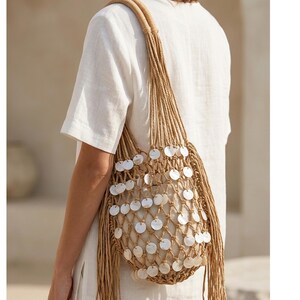 May include: A woven beige shoulder bag with a macrame design, adorned with white circular embellishments. The bag has a braided handle and long fringe detailing, creating a bohemian aesthetic. The bag is made of natural fibers.
