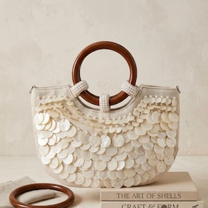 May include: A beige handbag featuring a crescent shape, adorned with layered white circular shell discs and small white beads. The bag includes a dark brown circular wooden handle and a matching detachable wooden ring accessory.