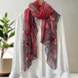 May include: A sheer, pink scarf with a paisley pattern in shades of pink, blue, and green is draped over a white button-down shirt. The scarf has a delicate, flowing appearance, and the shirt is neatly buttoned.
