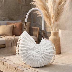 Könnte beinhalten: Weiß gehäkelte Handtasche mit Muscheldesign. Die Tasche hat einen geflochtenen weißen Griff und einen perlenbesetzten Schulterriemen. Goldfarbene Beschläge verbinden den Griff mit der Tasche. Die Tasche liegt auf einer Holzoberfläche.