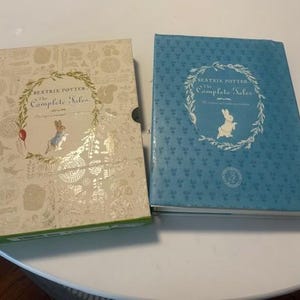 May include: Two copies of "Beatrix Potter The Complete Tales" are shown. One book is beige with gold detailing, the other is blue. Both feature a rabbit illustration within a floral wreath. The books are hardback.