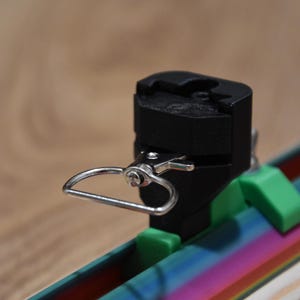 Könnte beinhalten: Eine schwarze und silberfarbene Metallklemme mit einem Scharnierring und grünen Stützen. Die Klemme ist an einer mehrfarbigen, regenbogenfarbenen Oberfläche befestigt. Der Hintergrund ist eine hellbraune Holzoberfläche.