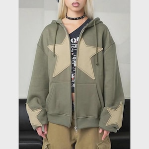 May include: Olive green zip-up hoodie with large tan star appliqués on the front and sleeves. The hoodie has a drawstring hood and a front zipper. The model is wearing a black top underneath and tan pants.