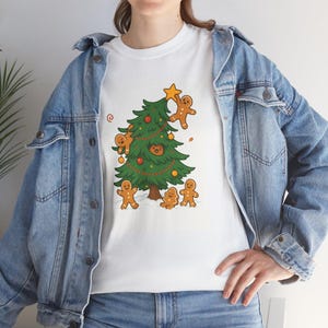 Könnte beinhalten: Weißes T-Shirt mit einem Weihnachtsbaum-Motiv, verziert mit Lebkuchenmännern und Ornamenten. Der Baum hat einen Stern an der Spitze und ist von Lebkuchenfiguren umgeben. Das Shirt wird mit einer blauen Jeansjacke getragen.