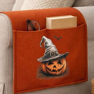 Könnte beinhalten: Ein orangefarbener Sofa-Armlehnen-Organizer aus Filz mit zwei Taschen, einer Brille und einem Buch. Die Vorderseite des Organizers zeigt einen grafischen Druck eines Kürbisses mit Hexenhut, akzentuiert durch eine kleine fliegende Fledermaus.
