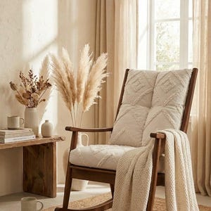May include: A wooden rocking chair with a cream-colored cushion and a matching throw blanket. The chair is placed on a woven rug, with a small wooden table to the left holding books, a mug, and a vase with dried flowers. The room has neutral tones.