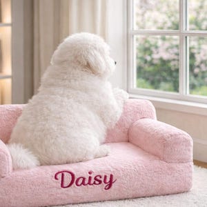 May include: A white dog sits on a pink pet sofa, gazing out of a window. The sofa is plush and has the name "Daisy" embroidered on it. The room features a bookshelf and a window, creating a cozy atmosphere.