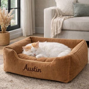 May include: A sleeping white and orange cat rests in a brown corduroy pet bed. The bed has a rectangular shape with raised sides and the name "Austin" embroidered on the front. The bed is on a neutral-colored rug.