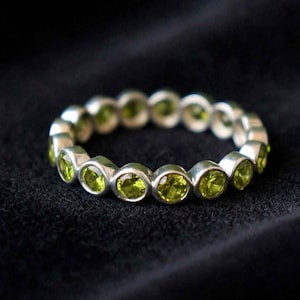 Könnte beinhalten: Ein silberner Eternity-Ring mit einer durchgehenden Reihe aus runden, facettierten, lindgrünen Peridot-Edelsteinen, die in einzelnen kreisförmigen Metallfassungen rund um den gesamten Umfang des Rings eingefasst sind.