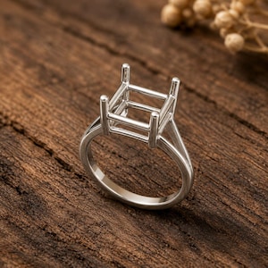 Könnte beinhalten: Ein silberner Ringrohling mit quadratischer Oberseite, zur Aufnahme eines Edelsteins. Der Ring hat ein klassisches Band und eine detaillierte Fassung, ideal für die Schmuckherstellung. Der Ring wird auf einer rustikalen Holzoberfläche präsentiert.