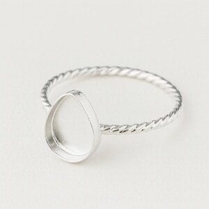 Könnte beinhalten: Ein silberner Ring mit einer tropfenförmigen Lünettenfassung. Das Ringband hat ein gedrehtes Seildesign. Die Fassung ist leer, bereit für einen Edelstein oder Harz. Der Ring wird auf weißem Hintergrund gezeigt.