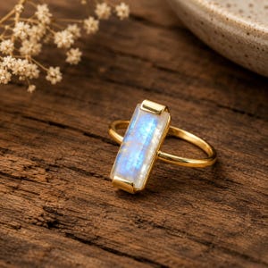 Könnte beinhalten: Ein goldener Ring mit einem rechteckigen, vertikal ausgerichteten Mondstein, der einen durchscheinenden, irisierenden blauen Schimmer aufweist. Der Edelstein ist in einer schlichten Metallfassung mit einem dünnen, polierten Goldring befestigt.