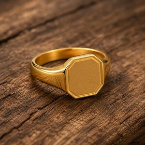 May include: A gold-toned signet ring featuring a smooth, flat, octagonal face. The ring has a polished, reflective surface and a solid band design. The metal has a warm, yellow hue, and the octagonal top is clearly defined with beveled edges.