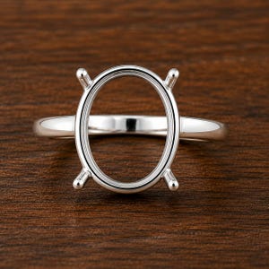 Könnte beinhalten: Eine silberne Ringfassung mit ovalem Mittelteil und vier Krappen. Der Ring liegt auf einer dunkelbraunen Holzoberfläche und zeigt seine polierte Metalloberfläche. Der Ring ist für einen Edelstein gedacht.