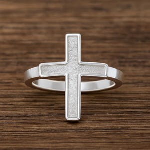 May include: A silver-colored ring featuring a textured cross design. The cross is the focal point, with a detailed surface. The ring band is smooth and simple, creating a contrast with the cross. The ring is set against a wooden background.