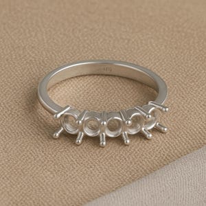 Könnte beinhalten: Ein silberner Ring mit fünf Fassungen für Steine. Der Ring besteht aus glänzendem Metall und hat ein schlichtes Band. Die Fassungen sind rund und haben kleine Zinken, um die Steine zu halten. Der Ring befindet sich auf einem neutralen Hintergrund.