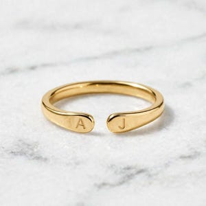 Puede incluir: Un anillo abierto de tono dorado con un acabado liso y pulido. Los dos extremos de la banda están ligeramente aplanados y redondeados, con la letra A grabada en un lado y la letra J grabada en el otro lado de la abertura.