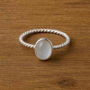 Könnte beinhalten: Ein silberner Ring mit einem gedrehten Band und einem ovalen, hellfarbenen Stein. Der Ring liegt auf einer Holzoberfläche, die sein Design und seine Handwerkskunst hervorhebt. Der Stein ist durchscheinend.