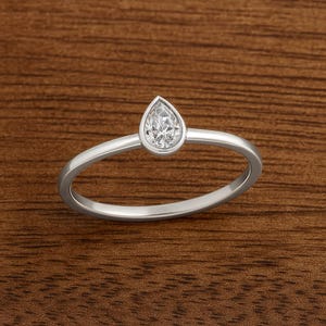 May include: A silver ring featuring a pear-cut, clear gemstone. The gemstone is set in a bezel setting, and the band is thin and polished. The ring is displayed on a wooden surface.
