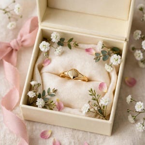 Puede incluir: Un anillo dorado con un diseño en forma de concha centrado en la banda, colocado dentro de una caja de color crema forrada con tela suave y decorada con pequeñas flores blancas y pétalos de flores rosas esparcidos.