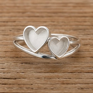 Könnte beinhalten: Ein silberner Ring mit zwei herzförmigen Fassungen. Der Ring hat ein Doppelband-Design, wobei die Herzen nebeneinander liegen. Die Herzen haben eine strukturierte, matte Oberfläche. Der Ring ist vor einem Holzgrund platziert.