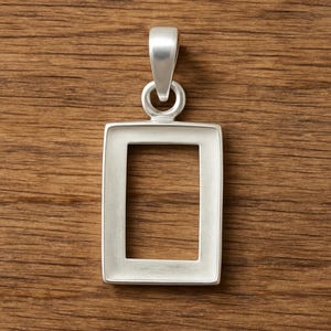 Puede incluir: Un colgante rectangular plateado con un lazo para una cadena. El colgante tiene un diseño simple y moderno con un centro hueco. El colgante se muestra sobre una superficie de madera.