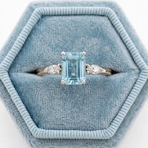 Könnte beinhalten: Ein Ring mit einem großen, rechteckigen, smaragdgeschliffenen hellblauen Edelstein in einer Metallschiene. Der Mittelstein ist auf jeder Seite von zwei kleineren, birnenförmigen, klaren Steinen flankiert, was ein symmetrisches Design mit polierter Oberfläche ergibt.