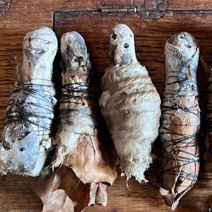 Può includere: Quattro piccole figure avvolte con superfici invecchiate e strutturate. Ognuna è legata con spago scuro e adornata con foglie secche. Le figure poggiano su una superficie di legno, evocando un'estetica rustica e artigianale.