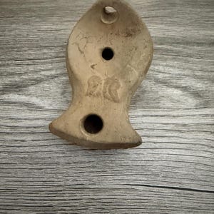 May include: A beige, fish-shaped ceramic oil lamp with a matte, textured surface. The object features three circular openings: one at the top, one in the center, and one at the base. There are faint, embossed markings visible on the central body.