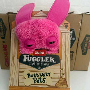 Könnte beinhalten: Ein pinkfarbenes Fuggler-Plüschtier mit großen Ohren und schläfrigen Augen schaut aus seiner Box. Die Box zeigt das Fuggler-Logo und die Worte "Funny Ugly Monster" und "Butt Ugly Pets."