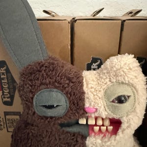 Op de afbeelding: Een pluchen konijn met een gespleten ontwerp. De ene kant is bruin, de andere is crèmekleurig. Het heeft een grijze oor, een gestikt oog en een mond met zichtbare tanden. Het speelgoed heeft een unieke, enigszins verontrustende esthetiek.