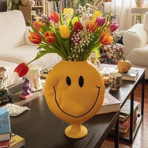 May include: A yellow smiley face vase filled with colorful tulips sits on a dark wood coffee table. The vase is the focal point of a cozy living room with a white sofa, patterned wallpaper, and various decorative items.