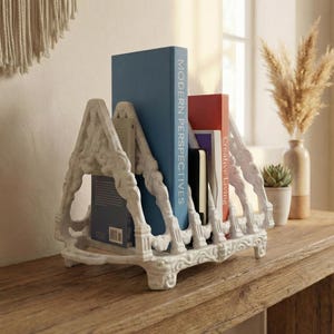 May include: A white, ornate book holder with a decorative design, holding several books with blue and red covers. The books are on a wooden table, with a window and decorative items in the background. The words "Modern Perspectives" and "Creative Living" are visible on the book spines.