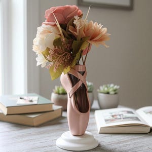 Könnte beinhalten: Eine rosa Ballettschuh-Vase mit einem Blumenstrauß in Rosa- und Pfirsichtönen. Die Vase steht auf einem weißen Sockel. Bücher und kleine Topfpflanzen im Hintergrund schaffen eine zarte, feminine Atmosphäre.