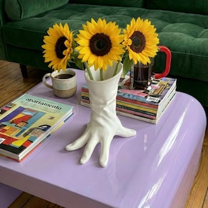 Puede incluir: Un jarrón blanco con forma de mano que contiene tres girasoles con pétalos amarillos y centros marrón oscuro. El jarrón está sobre una mesa de centro morada brillante, con un sofá de terciopelo verde al fondo. Una taza de café y revistas también están sobre la mesa.