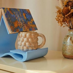 Puede incluir: Un atril azul claro con un libro titulado "The Art of Looking" y una taza beige con un patrón de burbujas. La taza se apoya en una bandeja azul claro. Un jarrón con flores secas está en el fondo.